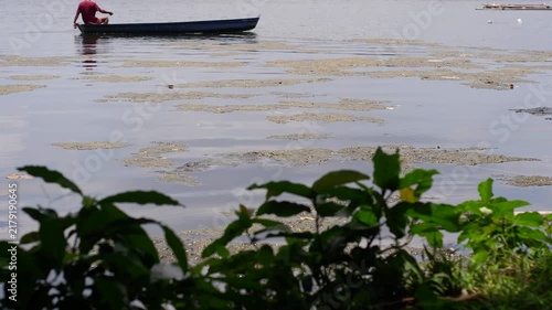 person in a small boat on a lake with dark, organic scum floating on the water's surface near the shore, where fish cages and structures are visible in the background.