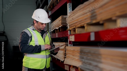Warehouse worker uses a digital tablet to check inventory and stored materials on industrial shelving during stock control and warehouse inspection.