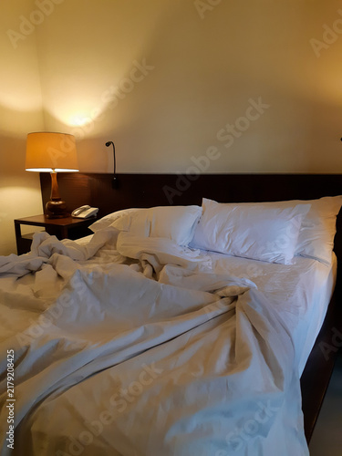 Messy unmade bed in a dark room with warm yellow light illuminating the wooden headboard and nightstand