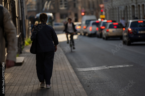 Konstfotografi A pedestrian in dark clothing moves along a city sidewalk while a cyclist threads past a queue of cars, the low sun stretching shadows over the pavement
