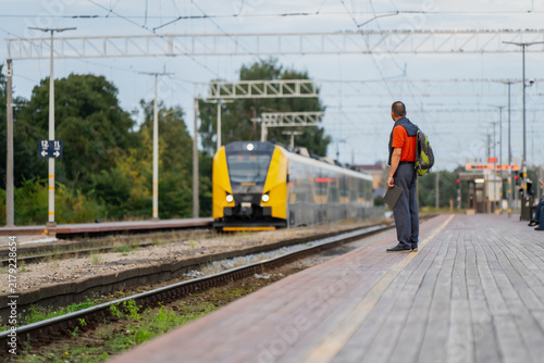 Regional rail stops depend on infrequent but reliable service, a single passenger waiting alone as a yellow train approaches under a web of overhead wires.
