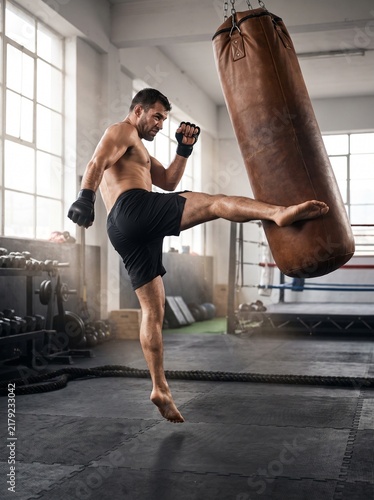 Dynamic muscular shirtless MMA fighter performing a jumping kick on a leather heavy bag in a bright gym