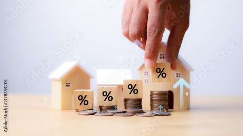 Hand placing percentage block on coin stack with wooden house models, real estate mortgage interest rate rise, property investment concept.