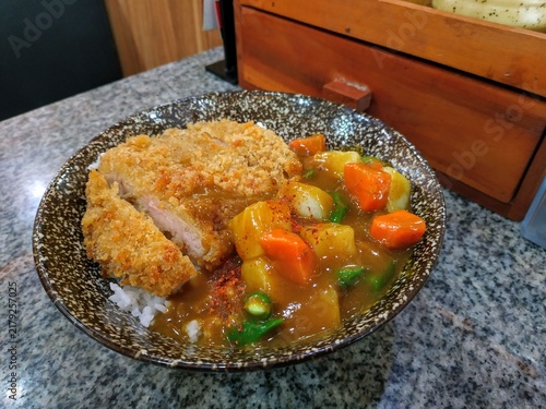Close-up view of authentic Japanese katsu curry rice served in a speckled ceramic bowl on a granite countertop. Features a crispy breaded chicken cutlet, rich brown curry sauce, chunky potatoes, carro