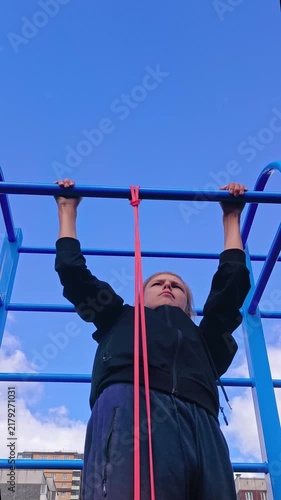 A teenager is hanging from an outdoor pull‑up bar using an elastic band, performing a workout to develop arm, back, and grip strength.
