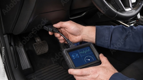 Mechanic plugging diagnostic scanner into obd port under car dashboard. Hands of man holding electronic diagnostic tool with screen showing car data. Computer diagnostics and repair of vehicle