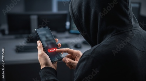 Rear view of an anonymous hacker in a black hoodie pressing a red hack button on a smartphone screen, mobile cybersecurity threat and cybercrime concept