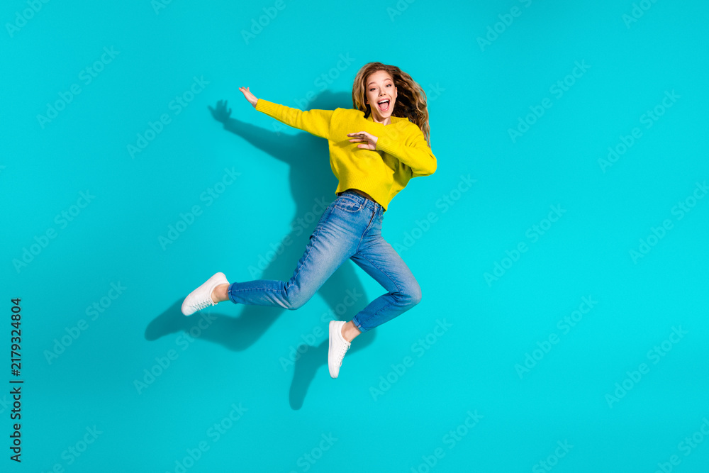 Fototapeta premium Young stylish woman wearing a yellow sweater jumps in the air against a blue background for a vibrant fashion lifestyle stock image