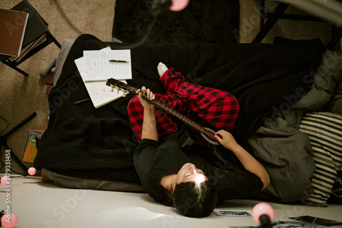 Bild auf Leinwand High angle view of young biracial woman playing acoustic guitar while lying on bed and writing song lyrics