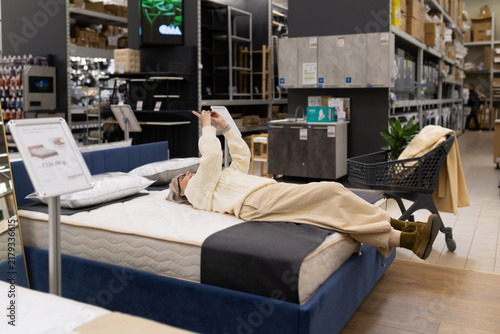 Woman lying on mattress, checking phone during furniture shopping