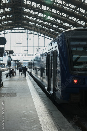 Passengers preparing to board a modern train at a historic railway station, framed by dramatic daylight through an expansive glass and steel roof.