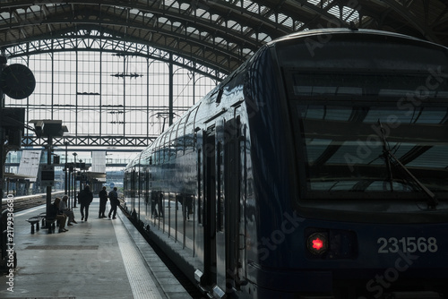 Passengers preparing to board a modern train at a historic railway station, framed by dramatic daylight through an expansive glass and steel roof.