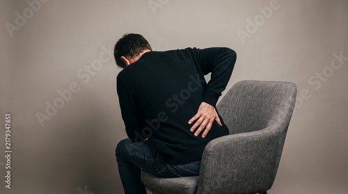 Man sitting on chair holding his lower back in pain, suffering from lumbar spine injury.
