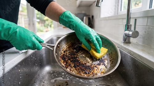 Cleaning a badly burned frying pan with a sponge and rubber gloves in a kitchen sink, showing stubborn burnt residue, dishwashing, household cleaning and kitchen hygiene