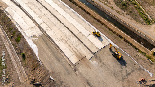 Aerial view of road construction site with concrete slabs being laid by a yellow road roller and an excavator near an irrigation ditch in Sleman, Special Region of Yogyakarta, Indonesia.
