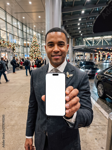 smiling african american man in a formal suit holding a smartphone with a blank white mockup screen in front of a building with christmas tree