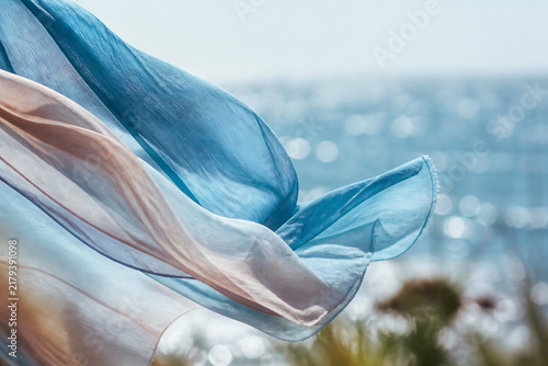 Flowing blue and pink fabric against a bright, hazy seascape with distant sparkling water.