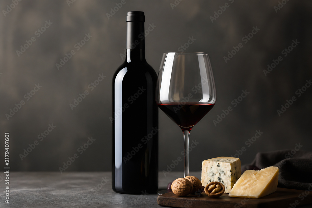 Obraz premium A bottle of red wine, a glass of red wine, cheese, and nuts are displayed on a dark surface against a moody background.
