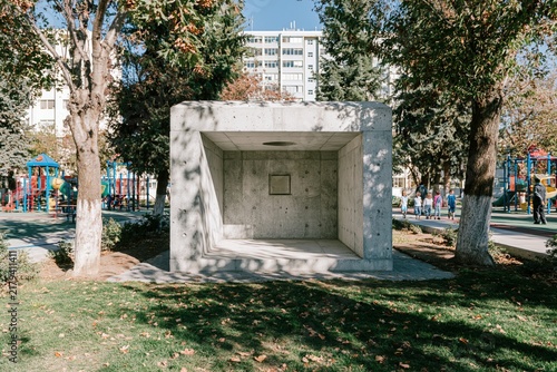Concrete protective shelter adjacent to a modern school campus featuring playground equipment, trees, and children playing in the background under bright daylight