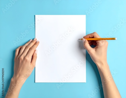 Top-down shot of hands holding a pencil above a blank white paper on a blue surface