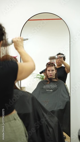 Hairdresser blow dries a hair section with a round brush, seen in a mirror reflection. Vertical salon scene for hair care, styling, beauty services and marketing