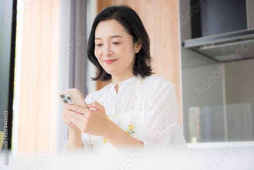 Obraz na płótnie A woman in her 50s smiles as she looks at her smartphone at home; she envisions a lifestyle where she enjoys browsing the internet and social media in between doing housework