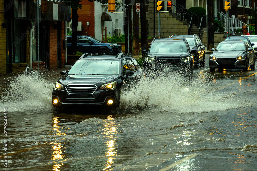 Obraz premium Vehicles drive through a flooded commercial street after a heavy rainstorm, splashing through standing water at dusk