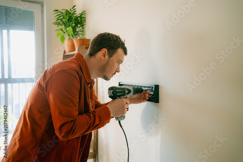 Man using a cordless drill to fasten a tv wall mount bracket onto a white wall, focusing on the installation process for a new television in a residential setting