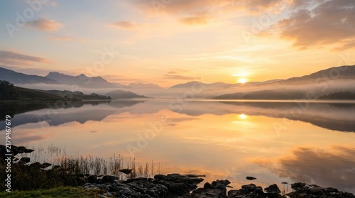 The sun rises over a serene lake, casting a warm golden glow across the water and surrounding mountains. Mist hangs low over the landscape, creating a tranquil and reflective atmosphere.
