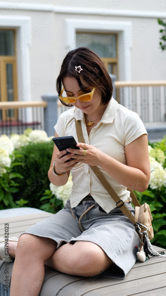 Obraz premium Young Woman Sitting Outdoors and Using Smartphone, Social Media, Text Messaging, Gen Z Digital Lifestyle, Leisure and Mobile Communication