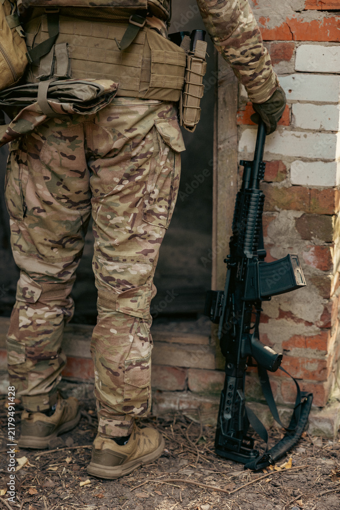 Obraz premium A soldier's legs next to an assault rifle. The soldier stands near the entrance to a ruined building