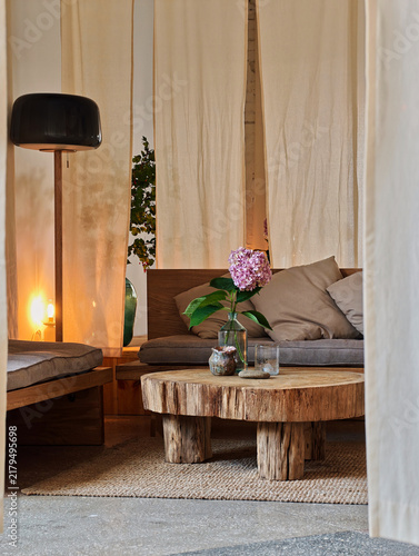 Aesthetic interior design of a tranquil living room with a raw log coffee table, fresh pink hydrangea in a glass vase, cozy low sofa cushions, elegant linen drapes, and warm floor lamp.