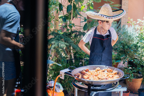 Картината върху платно Young boy learning outdoor cooking while watching paella simmer in sunny Mediter