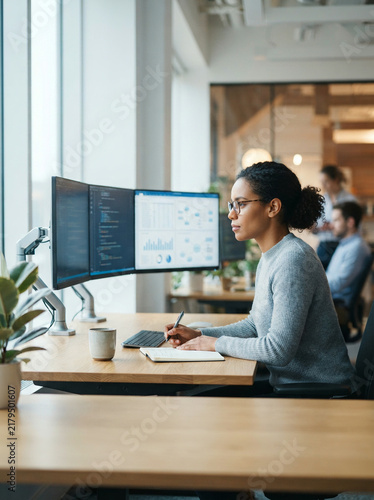 Woman writing in a notebook beside two monitors in an office in soft daylight