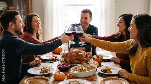 Happy multiethnic family and friends celebrating Thanksgiving dinner together, toasting with red wine glasses over roasted turkey and traditional autumn holiday food at home