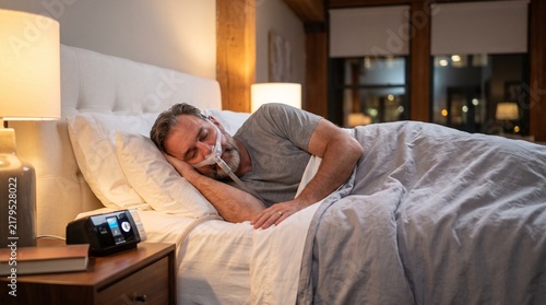 Middle aged man sleeping peacefully in a cozy bedroom using a CPAP machine and nasal mask for sleep apnea therapy at home.