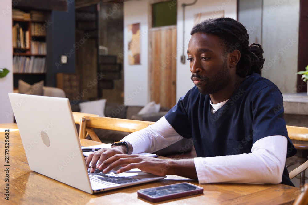 Obraz premium African man wearing scrubs working on laptop at wooden table at home, smartphone nearby, copy space