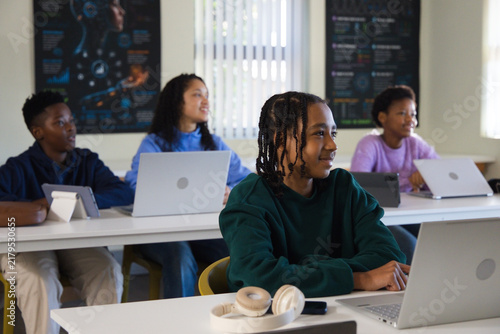 African American teenage classmates using laptops, tablet stand and white headphones in classroom