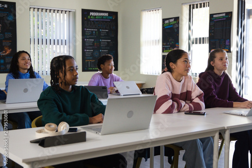 Diverse teenage students coding on silver laptops in coding lab with headphones and tablet posters