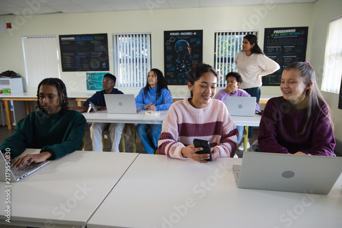 Diverse classmates using laptops and phone in computer lab with teacher supervising near posters