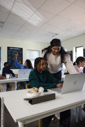 Indian teacher leaning over African American teen student, guiding on silver laptop in school