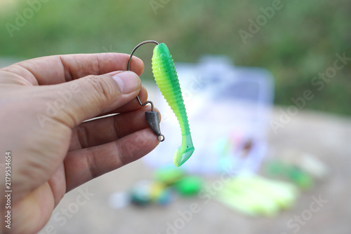 Fotografie Hand holding a bright green soft plastic paddle tail fishing lure with jig head and hook, featuring colorful fishing tackle in a blurred outdoor background