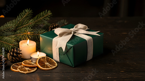 Festive Gift on Rustic Wood: A beautifully wrapped green gift with a cream ribbon sits beside flickering candles, pine cones, and dried orange slices on a dark wooden surface.