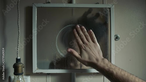 Hand wipes fogged bathroom mirror revealing face of young woman behind steam. Woman looking at reflection through wet glass after shower. Moisture condensation and dramatic mood concept