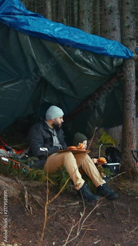 Man eating a sandwich at a makeshift survival shelter in the woods