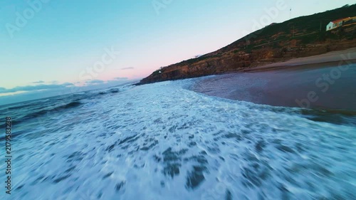 Soft waves wash warm sand at twilight. Sky blushes pink, fading to deep blue. Rocks jut sharp against crashing surf. Empty shore holds quiet, lonely beauty