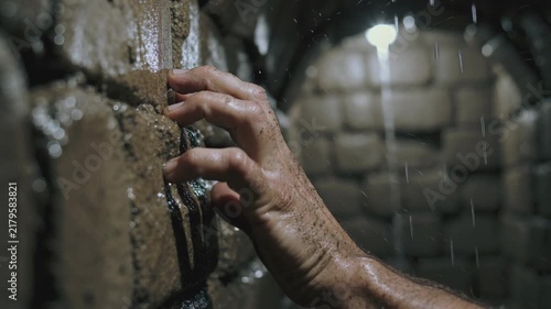 Wet hand of man touching brick wall in dark tunnel with dripping water. Closeup of dirty fingers sliding on stone wall in underground dungeon. Horror escape concept with rain falling in tunnel