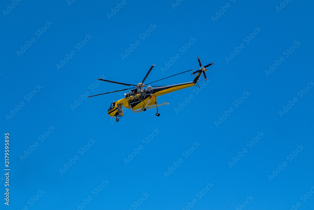 Fototapeta premium Yellow firefighting helicopter with water snorkel flying under blue sky on wildfire mission