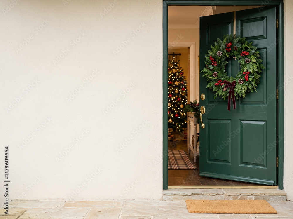 Fototapeta premium Open green front door decorated with a holiday wreath revealing a Christmas tree inside. Exterior home entrance with blank light wall providing copy space on the left