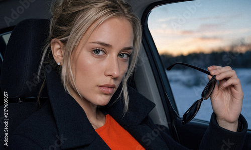 A young blonde woman sits inside a car holding sunglasses, staring directly at the camera with a confident, intense expression during dusk.